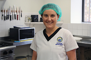 A woman wearing a hairnet stands in a kitchen. A microwave is on a bench behind her and a series of kitchen knives hangson a wall.