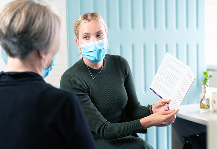 A woman wearing a surgical mask and holding a printed booklet speaks with another woman.