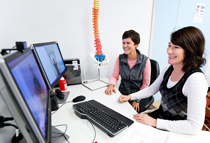 Two women site side by side at a desk in front of two monitors. A model of a spine and pelvis is behind on of the women.