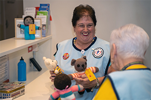 A woman wearing a tunic that has a badge reading Friends of Rockingham General Hospital holds some knitted toys