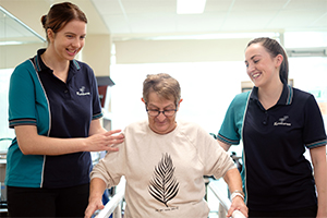 Two female physiotherapists stand either side of an older woman as she walks.