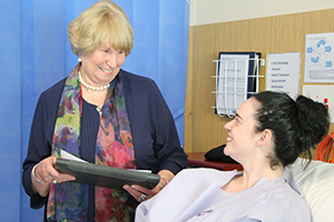 A woman holding some documents stands talking to a young woman in a hospital bed.