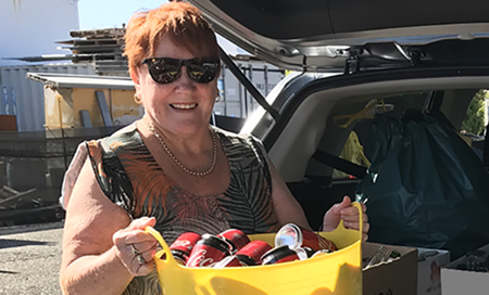 A woman wearing sunglasses stands beside the open boot of a car holding a crate of recyclable cans