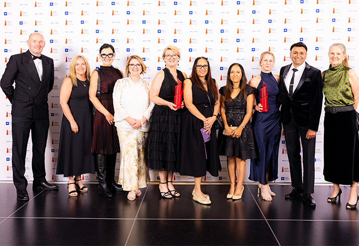ten people standing in front of a white banner with logos, two people are holding trophies